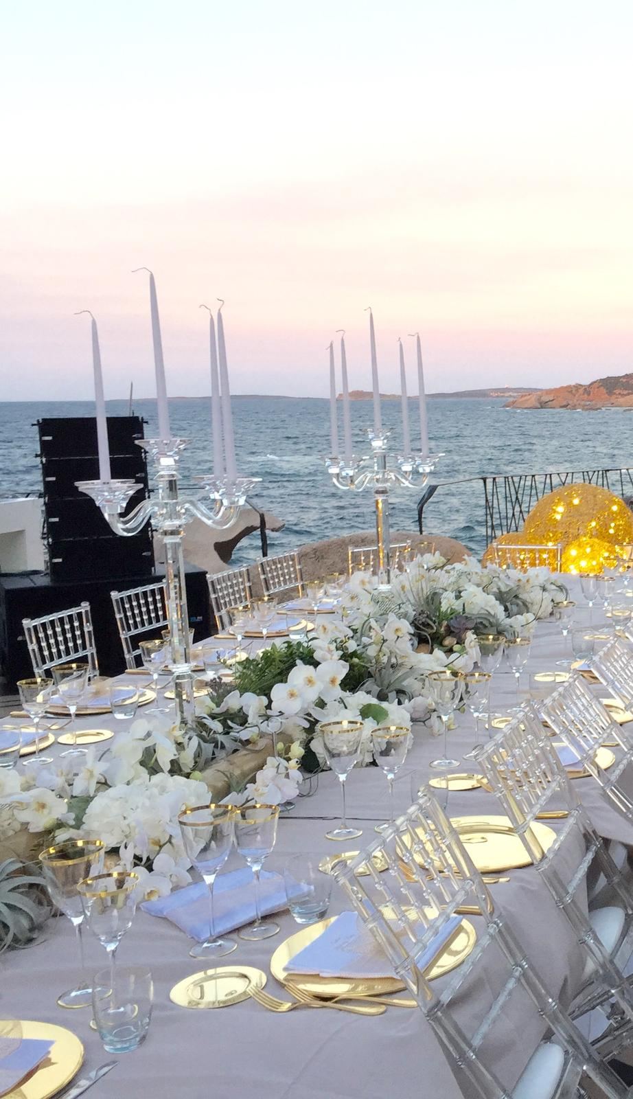 Tavolata al tramonto sul mare con candelabri e sedie trasparenti